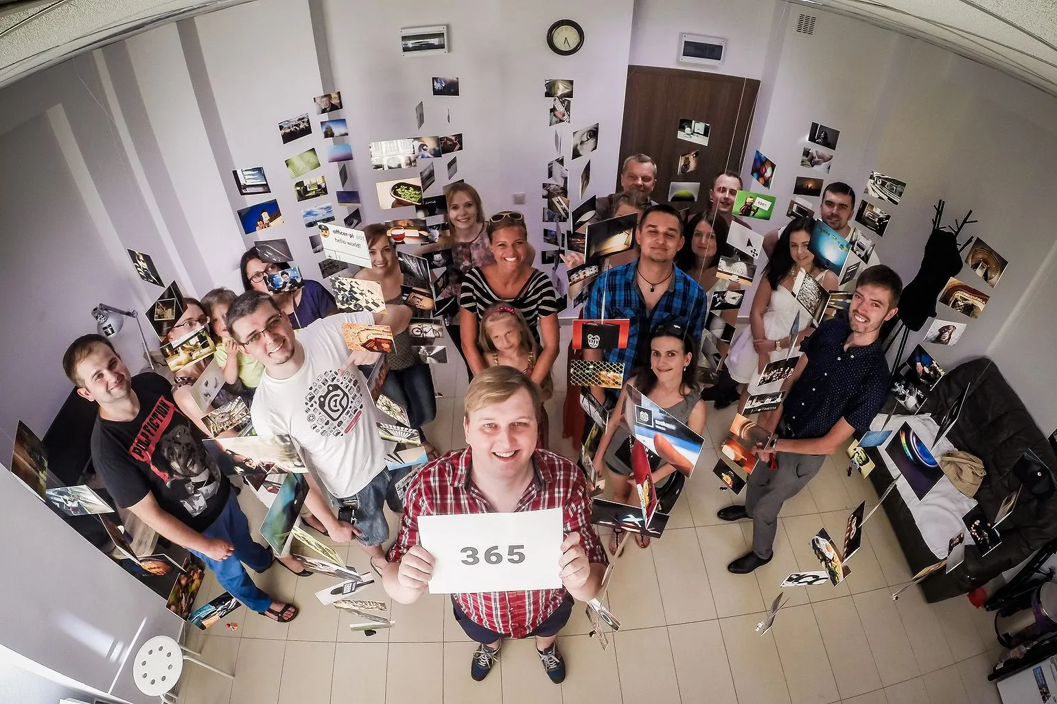 Wide-angle shot from above: a man in a checked shirt holds a sheet with the number 365, surrounded by a dozen people and photo prints hanging in the room
