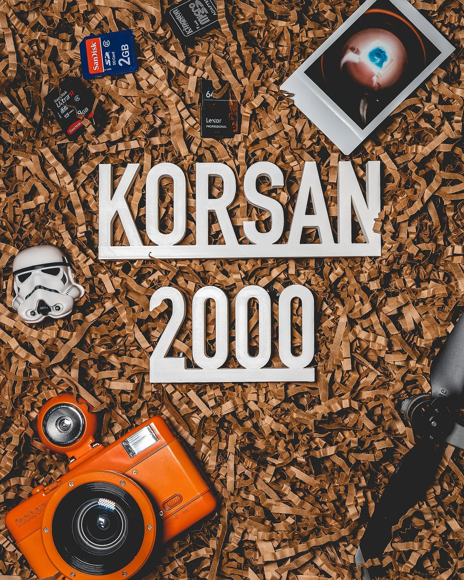 White letters reading KORSAN 2000 on brown shredded paper, surrounded by an orange camera, memory cards, a stormtrooper figure and an instant photo