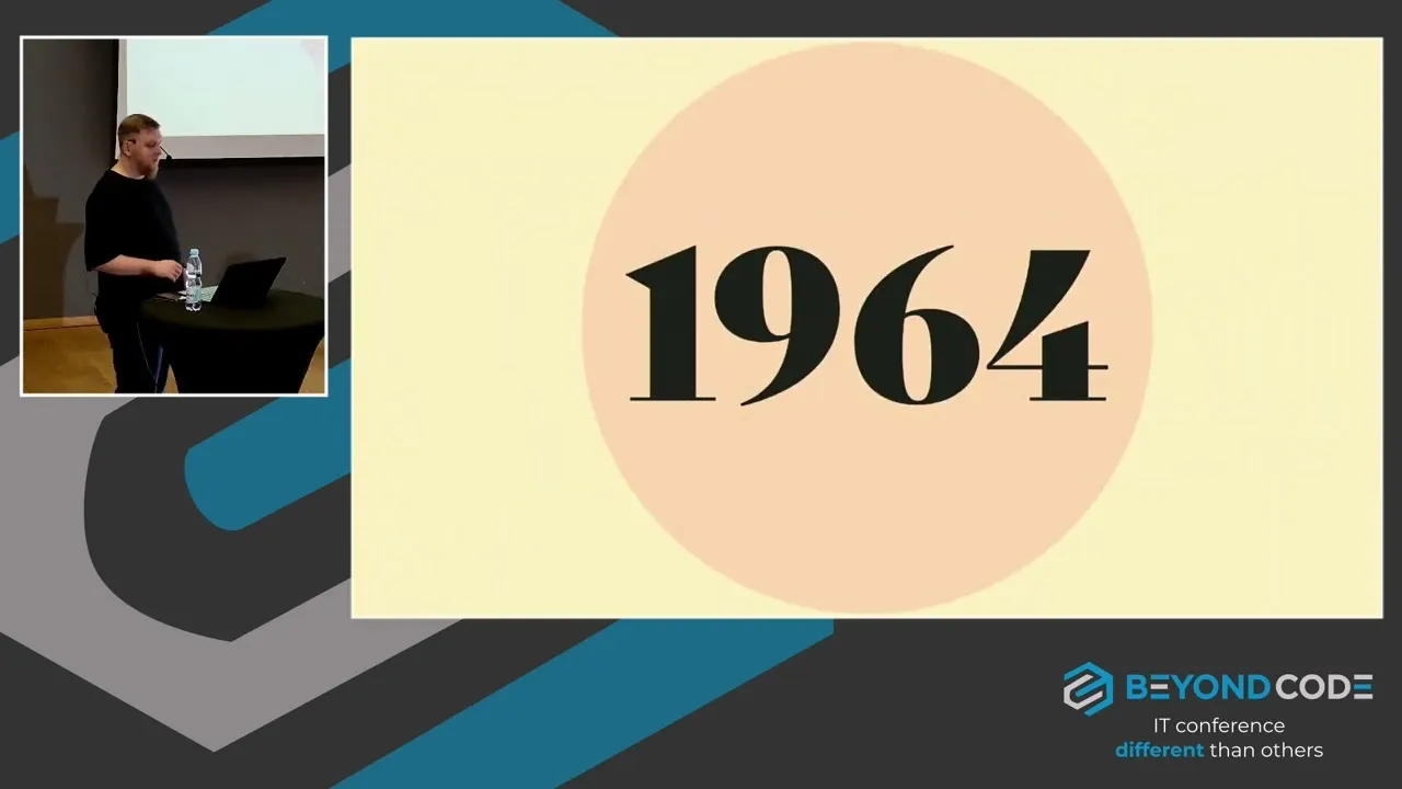 Slide showing the number 1964 on a beige circle, with a small inset of the speaker at a laptop on the Beyond Code stage