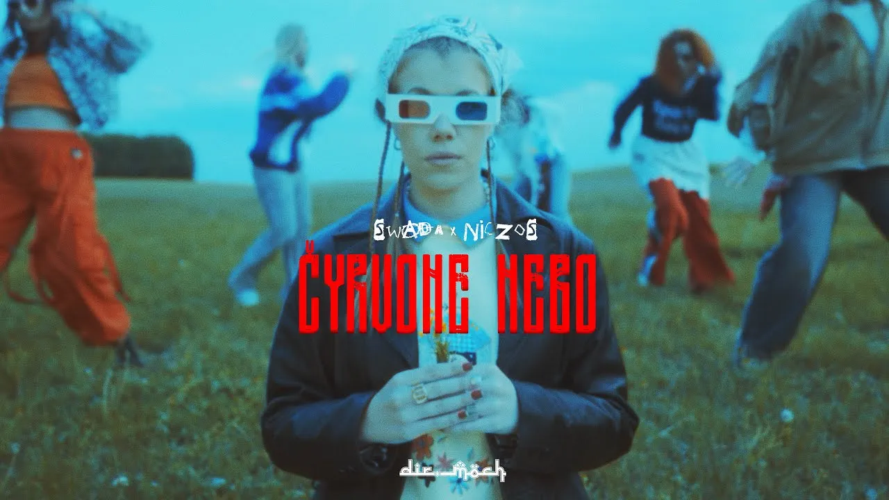 Still from the “Čyrvone Nebo” video: Niczos in 3D glasses in a meadow, people dancing behind her
