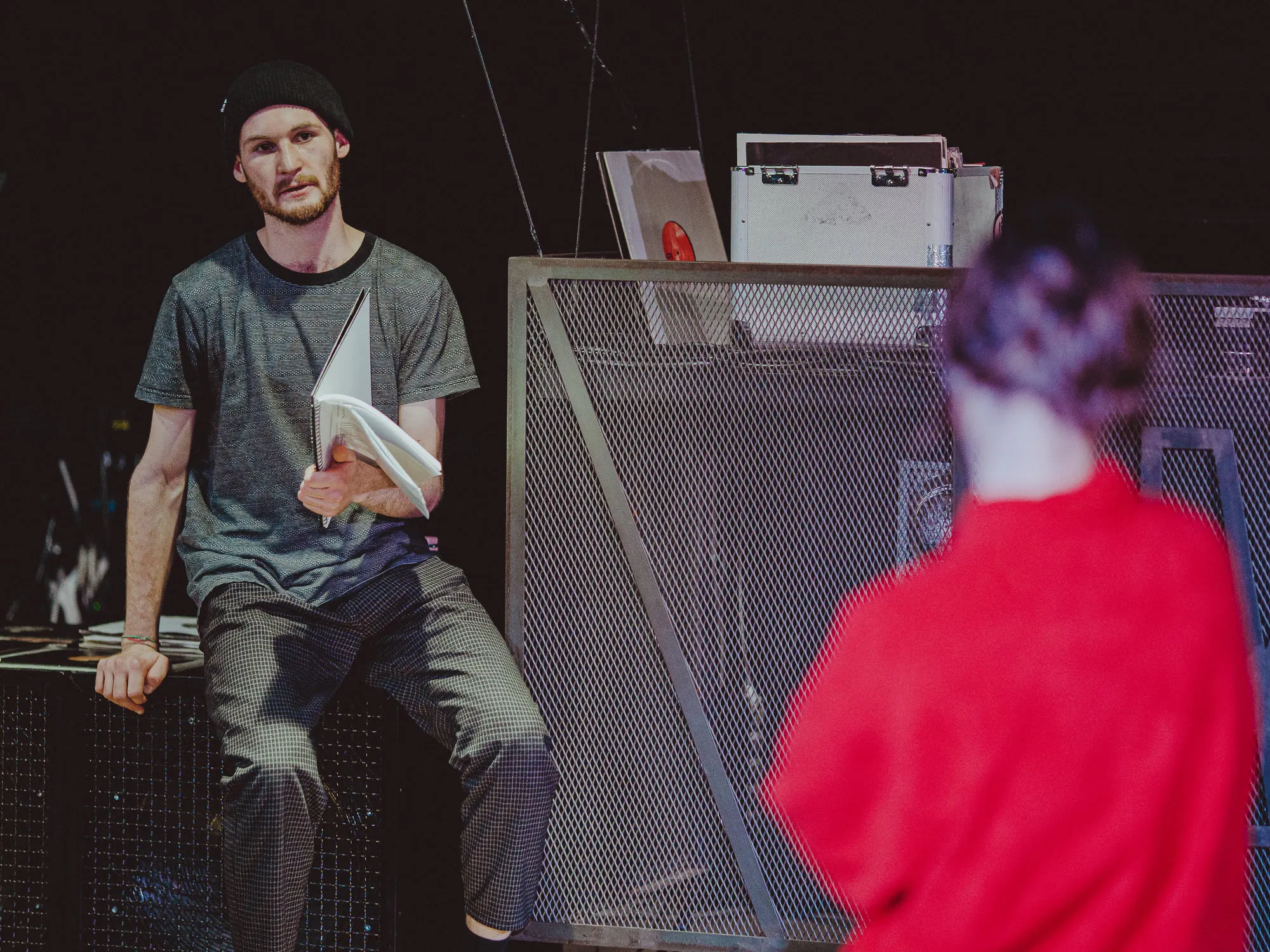 A young man in a beanie sitting with papers in hand next to a mesh cage with gear, a person in a red top in the foreground