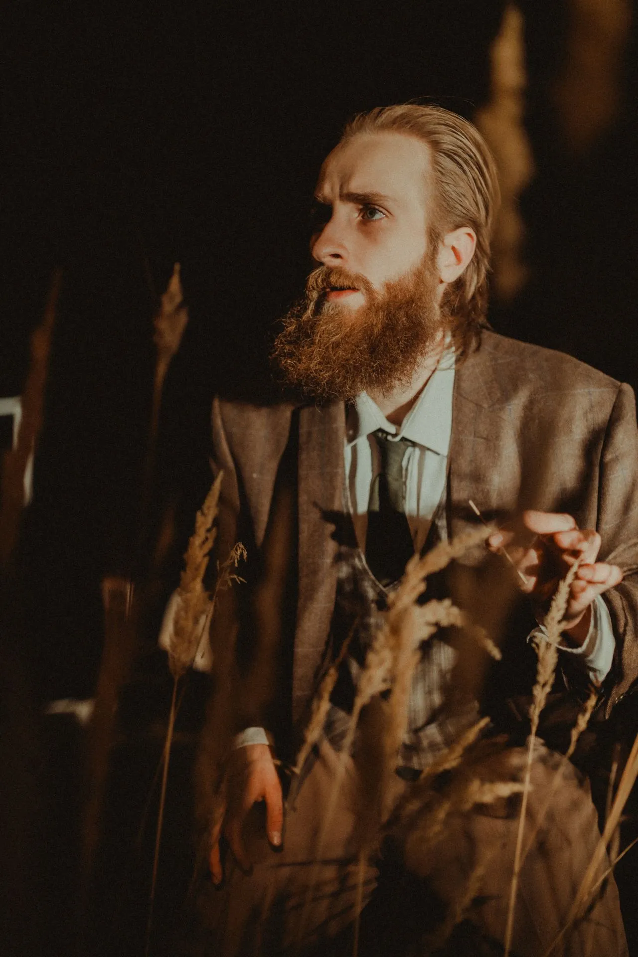 Marcin Tomkiel in a jacket and tie among tall dry grasses on a dark stage