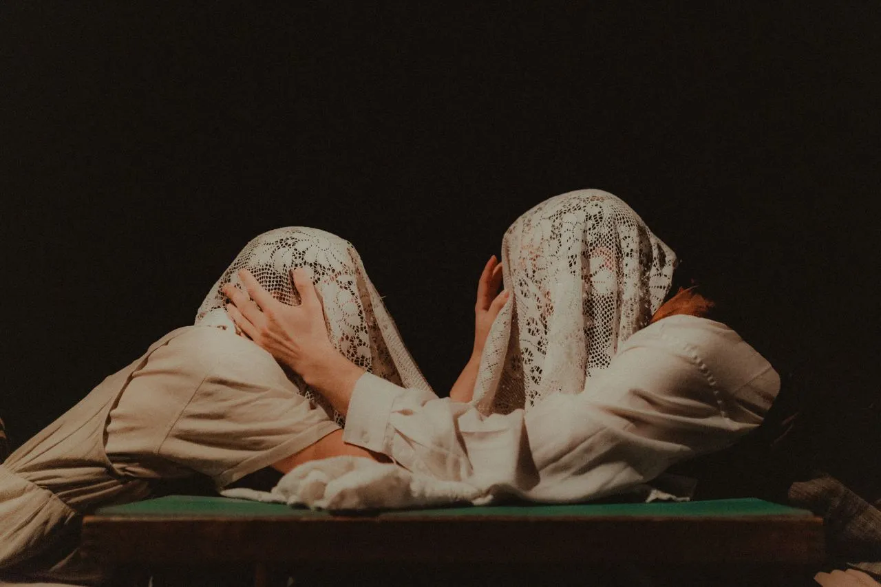 Two people leaning towards each other across a table, their heads covered with a lace tablecloth