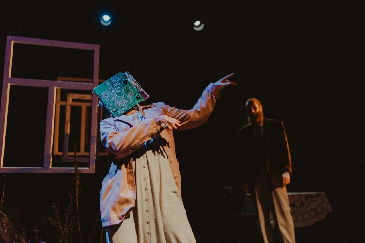 A performer gesturing with a sheet held over her face in front of empty window frames; Marcin Tomkiel stands in the background