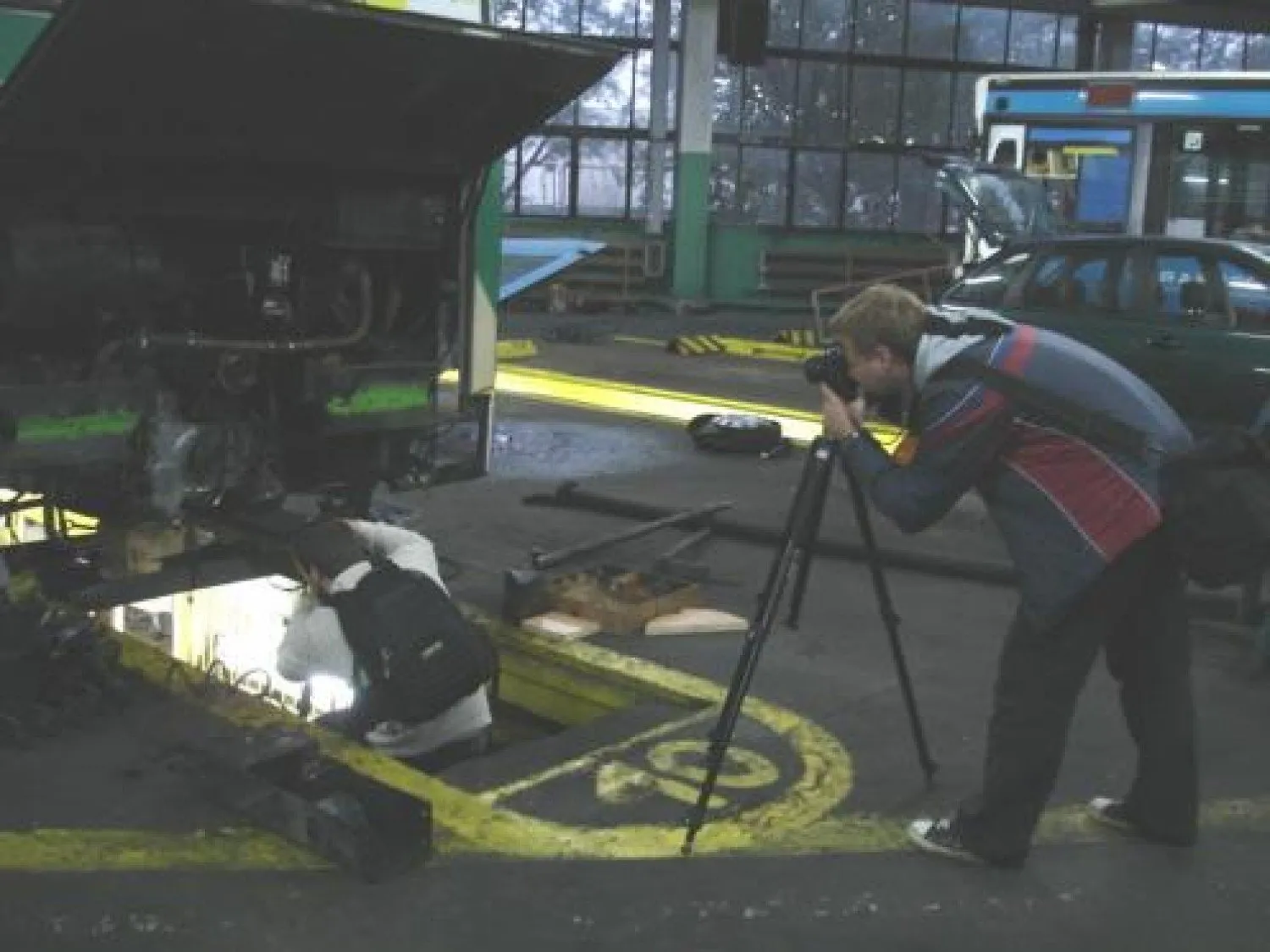 Two photographers in the KPKM repair hall, one shooting from the inspection pit under a bus, the other from a tripod