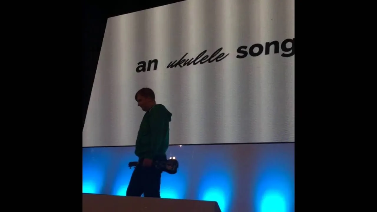 Silhouette of a man holding a ukulele on the dark Front-Trends stage, with “an ukulele song” on the screen behind him