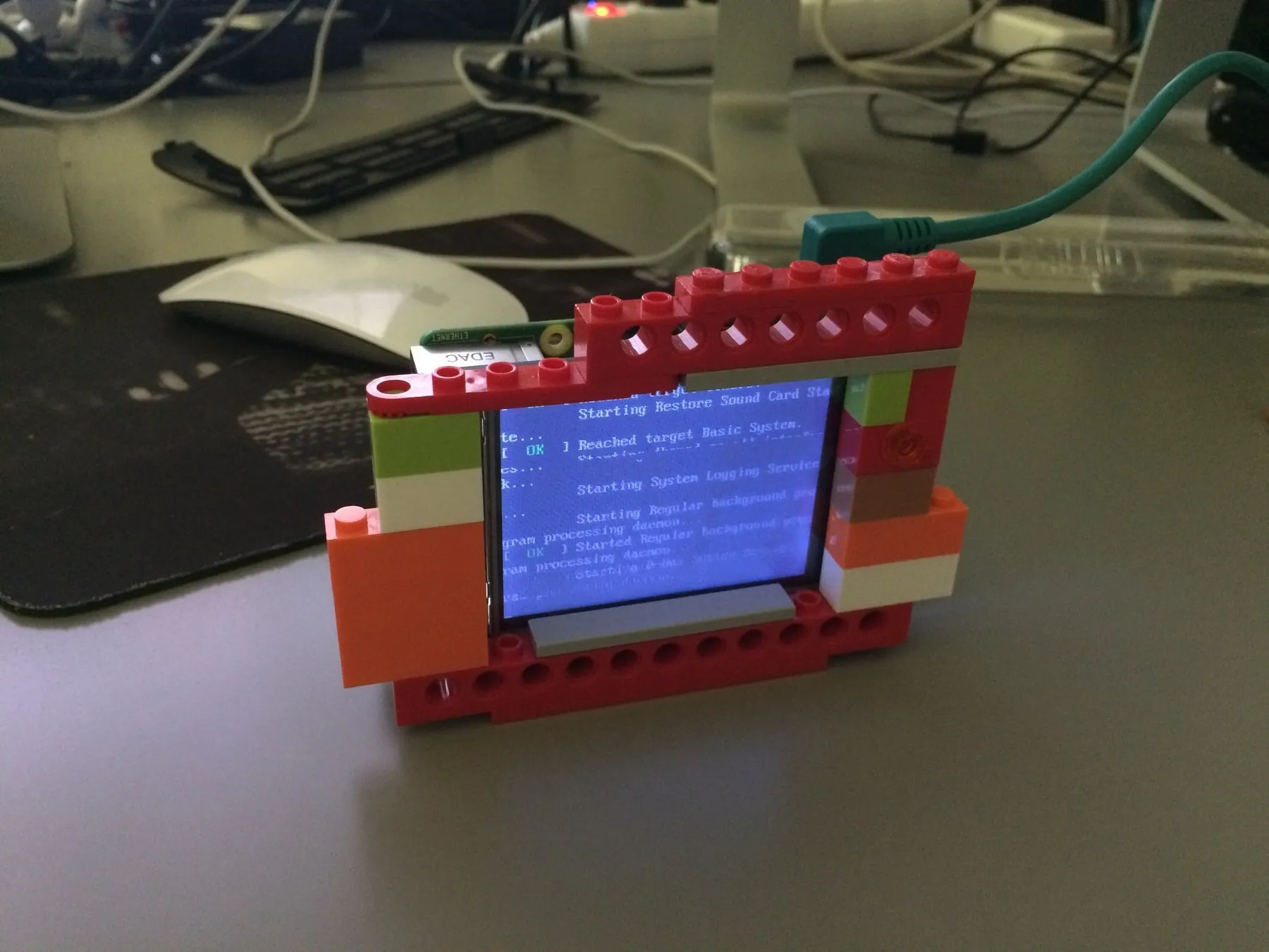 A small display in a frame of red and orange LEGO bricks on a desk, showing system boot text