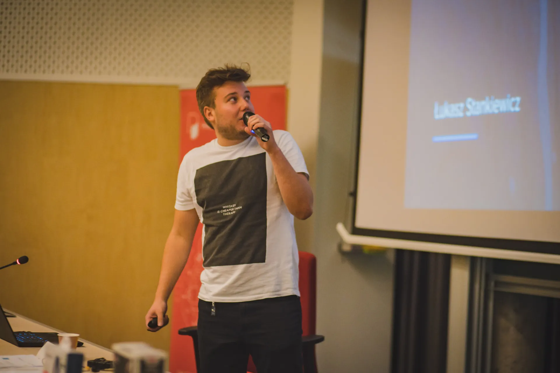 Speaker in a white T-shirt holding a microphone in front of a screen reading Łukasz Stankiewicz