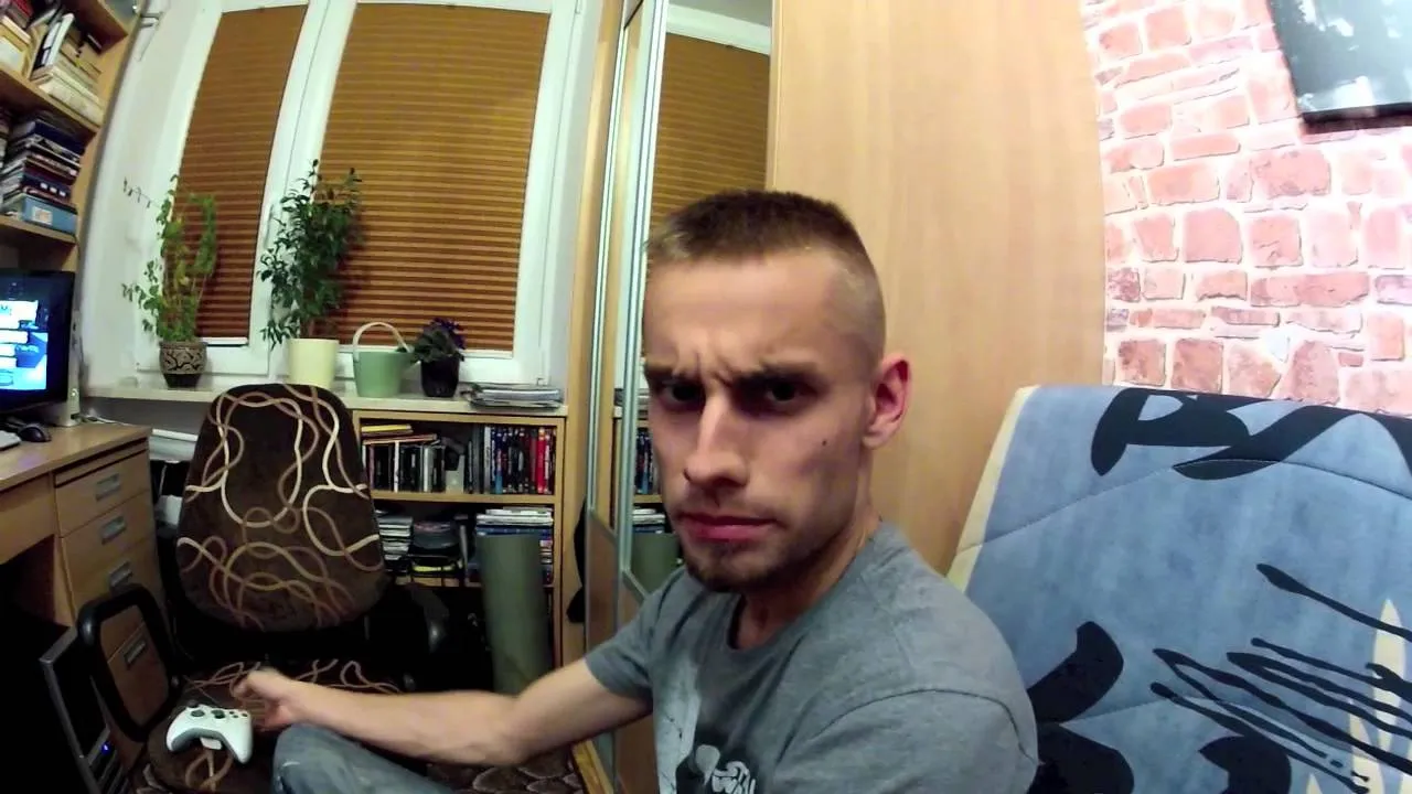 Film still: a young man in a grey T-shirt eyes the camera suspiciously, next to a desk with a monitor and a game controller
