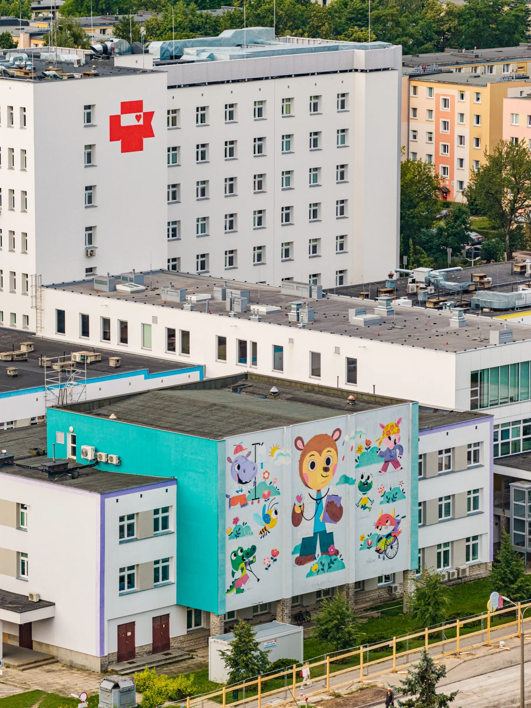 The Białystok children’s hospital from above in 2026: a low building with a turquoise side wall and the bear-doctor mural, behind it a white block marked with a red cross