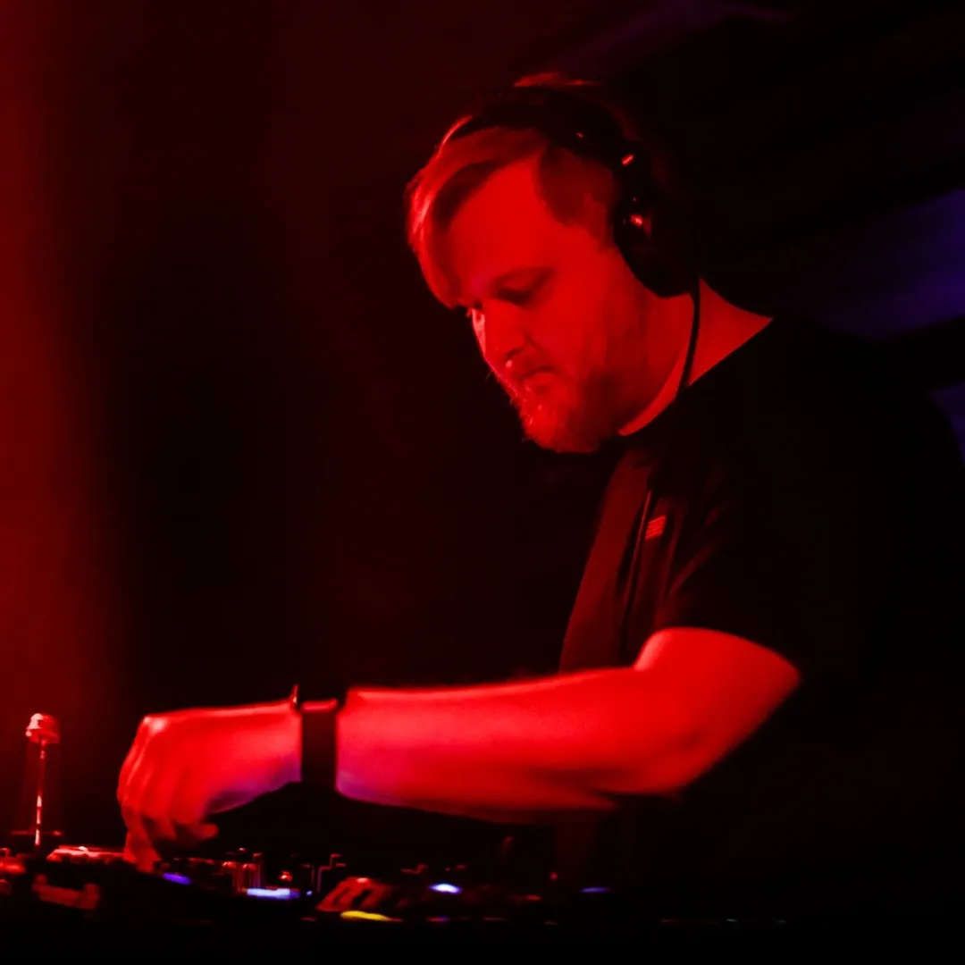 A man in headphones leaning over DJ gear, lit in red