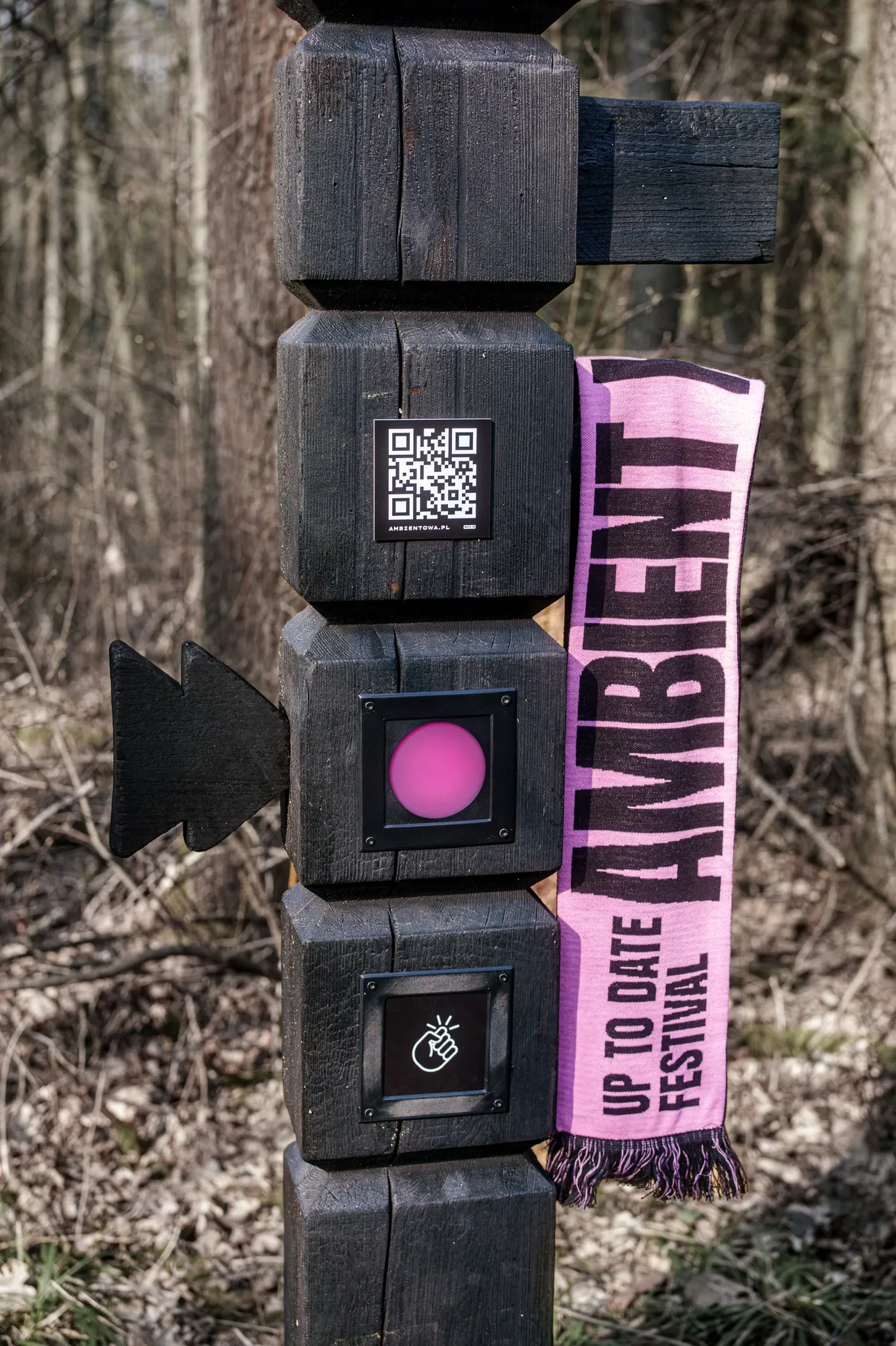 Part of a totem with a QR code, a glowing pink light panel and a knock pictogram; an Up To Date Festival scarf reading AMBIENT hangs beside it
