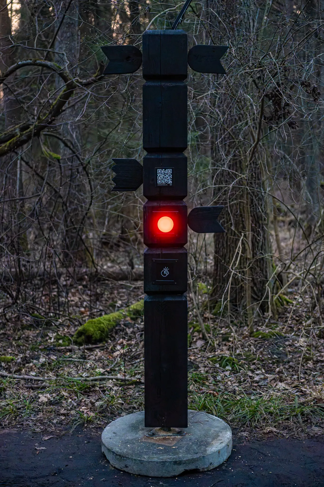 A totem at dusk with its red light on, standing on a concrete base next to a forest path