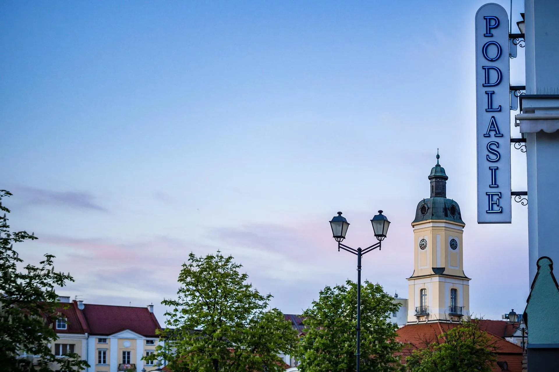 The tower of Białystok's town hall at dusk, with a street lamp, trees and a vertical sign reading Podlasie in the foreground