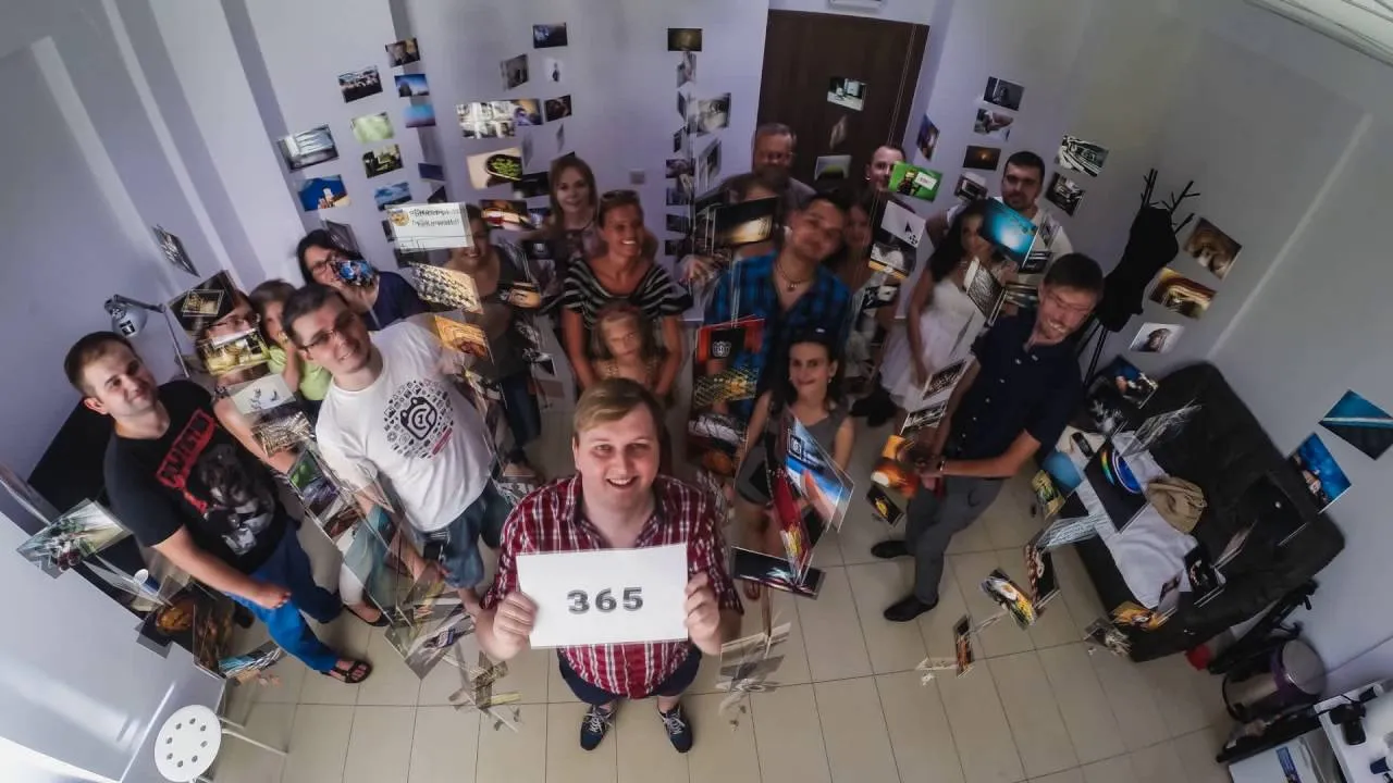 Still from the video “365 dni korsana”: a man holding a “365” sign among a dozen people and photo prints hanging in the room